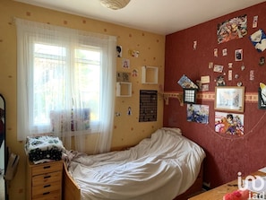 Une chambre à coucher comprenant un lit, une commode en bois, une fenêtre avec des rideaux légers et un mur décoré de divers tableaux et affiches.