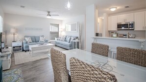 A modern living room with a sofa, coffee table, and dining table. There is a kitchen area with cabinets and a microwave. - Seas the Day Cottage (Gulfport)
