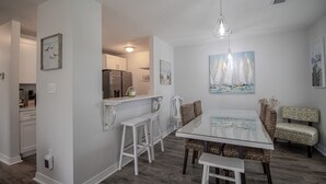 A dining area with a glass table and white chairs, a kitchen in the background, and a painting of sailboats on the wall. - Seas the Day Cottage (Gulfport)
