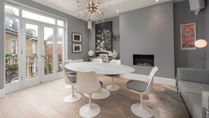A modern dining area with a round table and white chairs, a chandelier, and a fireplace. - Maida Vale Elegance (London)