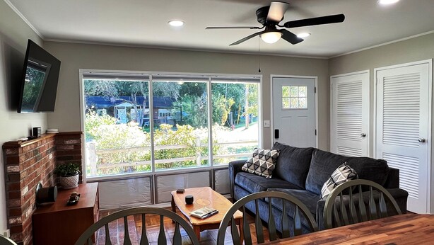 A living room with a sofa, a dining table, and chairs. There is a TV mounted on the wall, a ceiling fan, and a window with a view of the outside. - Welcome home in a welcoming town! (Littleton)