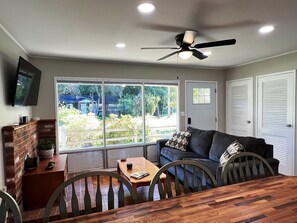 A living room with a sofa, a dining table, and chairs. There is a TV mounted on the wall, a ceiling fan, and a window with a view of the outside. - Welcome home in a welcoming town! (Littleton)