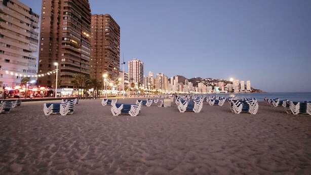 Una playa con filas de sillas y sombrillas, una orilla arenosa y edificios altos al fondo.