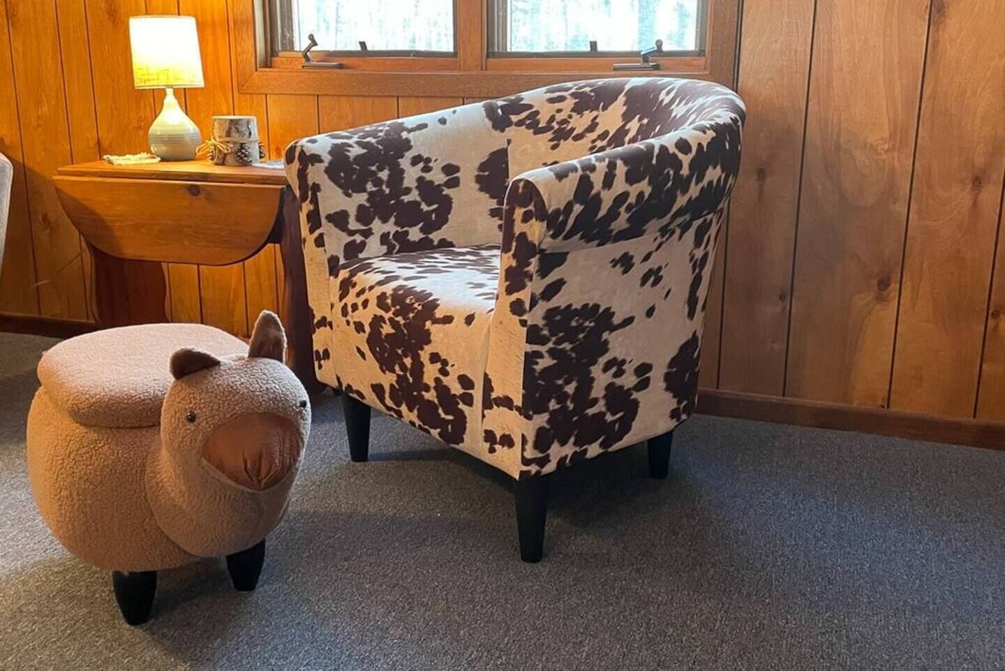 Une pièce avec un fauteuil à motifs de vaches, un petit pouf en forme d’agneau et une lampe posée sur une table d’appoint en bois.