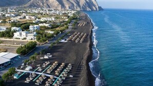 A coastal town with a beach lined with umbrellas and sun loungers, a mountain in the background, and white buildings. - Perissa Art Villa - Kitchen & Washing Machine (Santorini)