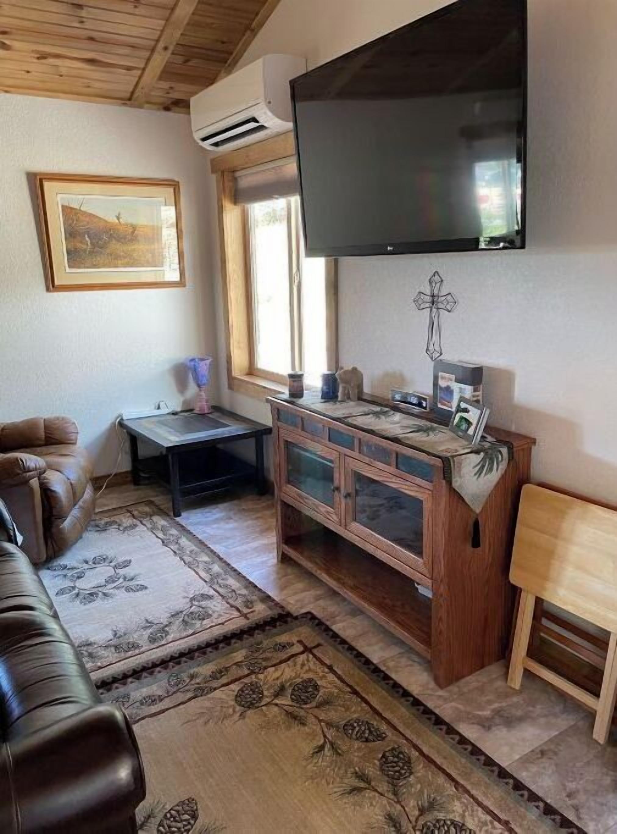 A living room with a wooden ceiling, a mounted TV, a cross on the wall, a sofa, a small table, and a cabinet.