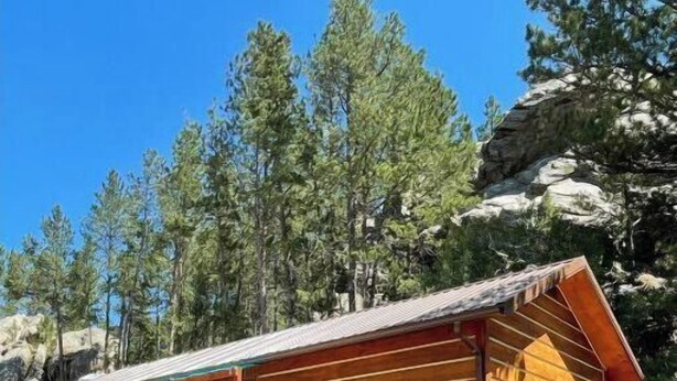 Una baita di legno con veranda, immersa tra alberi e terreno roccioso.