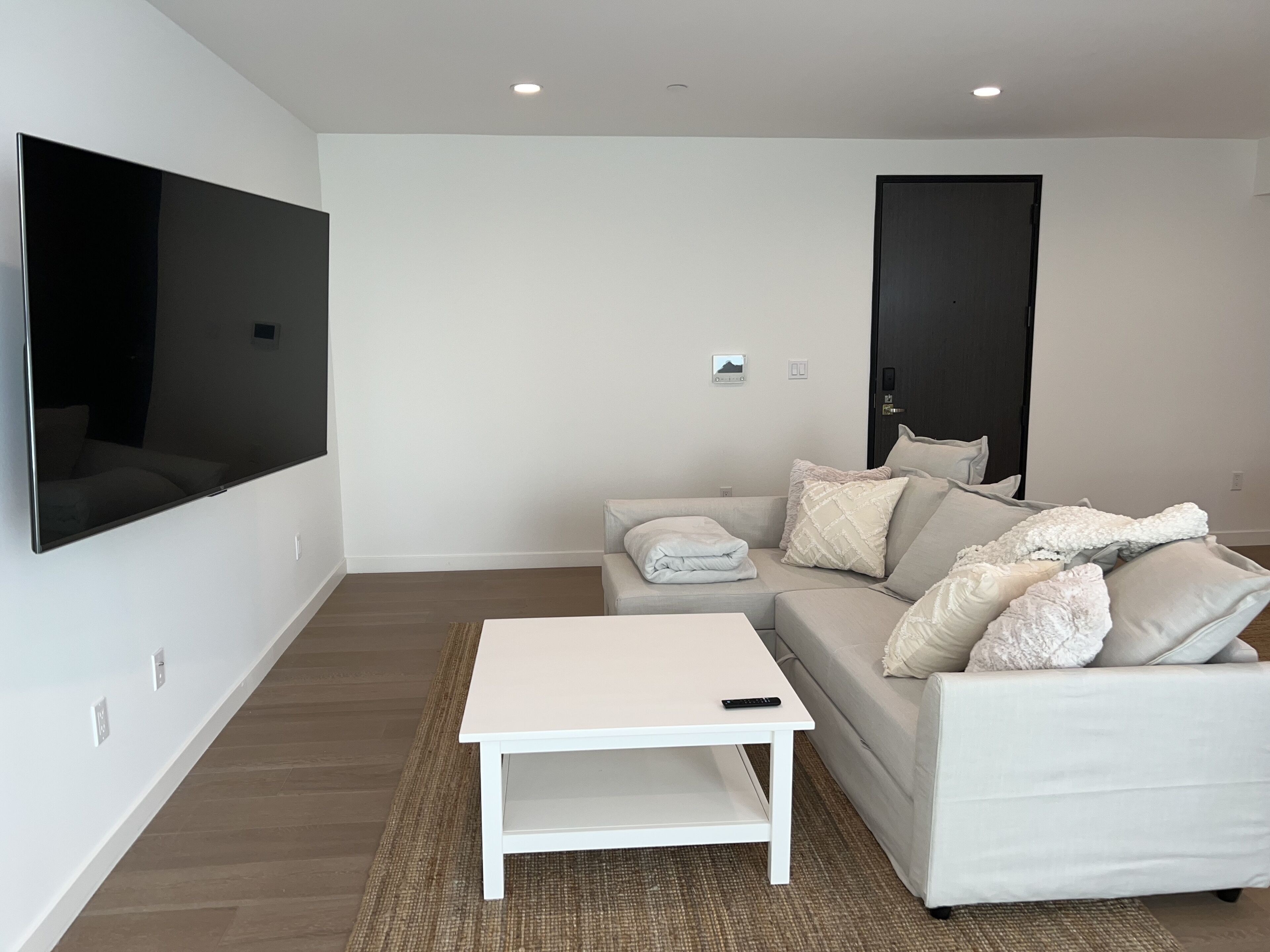 A modern living room with a flat-screen TV mounted on the wall, a white sofa with cushions, a white coffee table, and a door with a handle.
