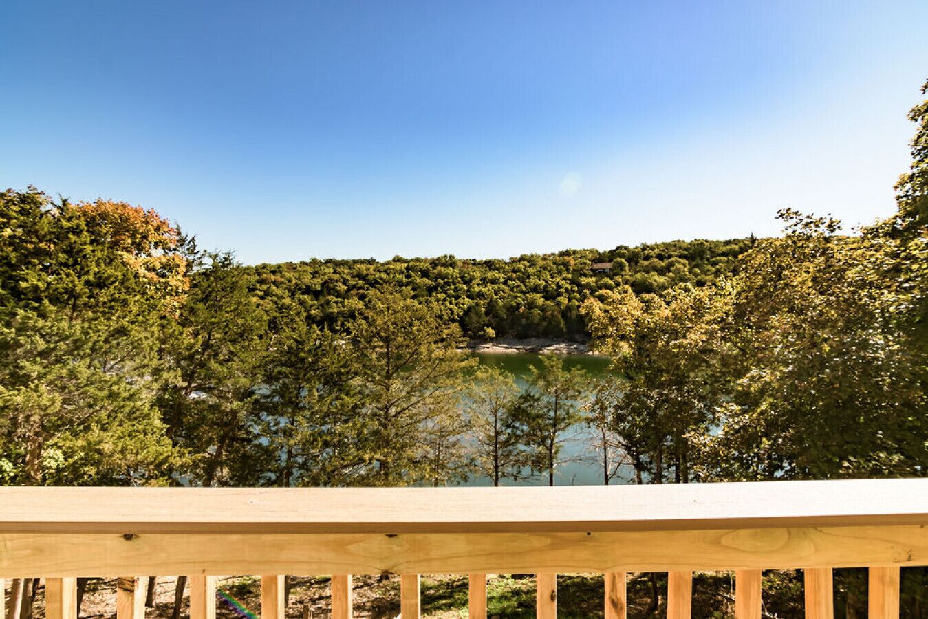 A wooden deck overlooking a lake with dense forest and a house on a hill in the background.
