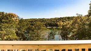 Una terraza de madera con vistas a un lago, rodeado de un bosque denso y con una casa en una colina al fondo.