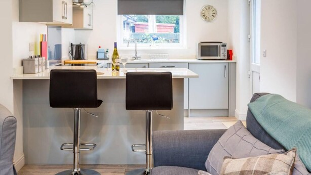 Eine kleine Küche mit Fenster, einer Mikrowelle und einer Wanduhr. Zwei Barhocker und ein graues Sofa mit Decke.