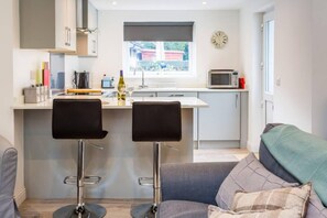 A compact kitchen with a window, a microwave, and a clock on the wall. There are two bar stools and a grey sofa with a blanket. - Ideal Couples Base for Exploring the Isle of Arran (Isle of Arran)