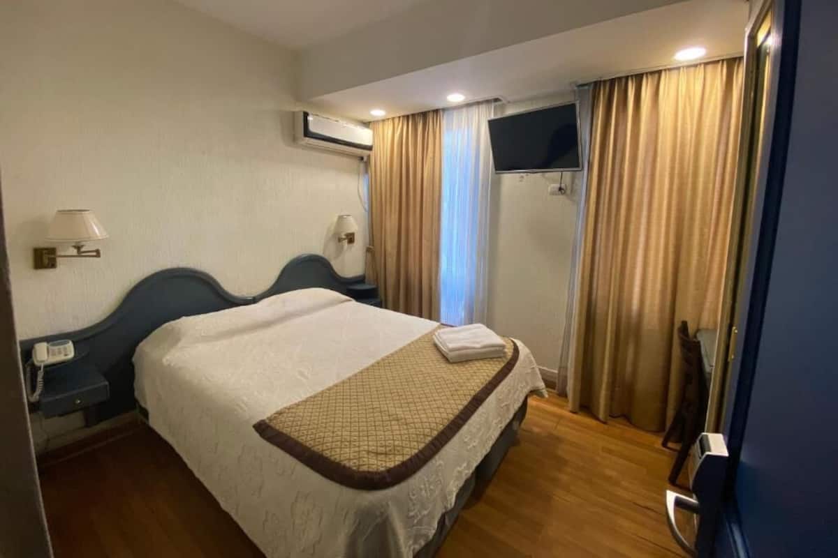 A hotel room with a bed, a TV, curtains, a bedside table with a lamp, and an air conditioning unit.