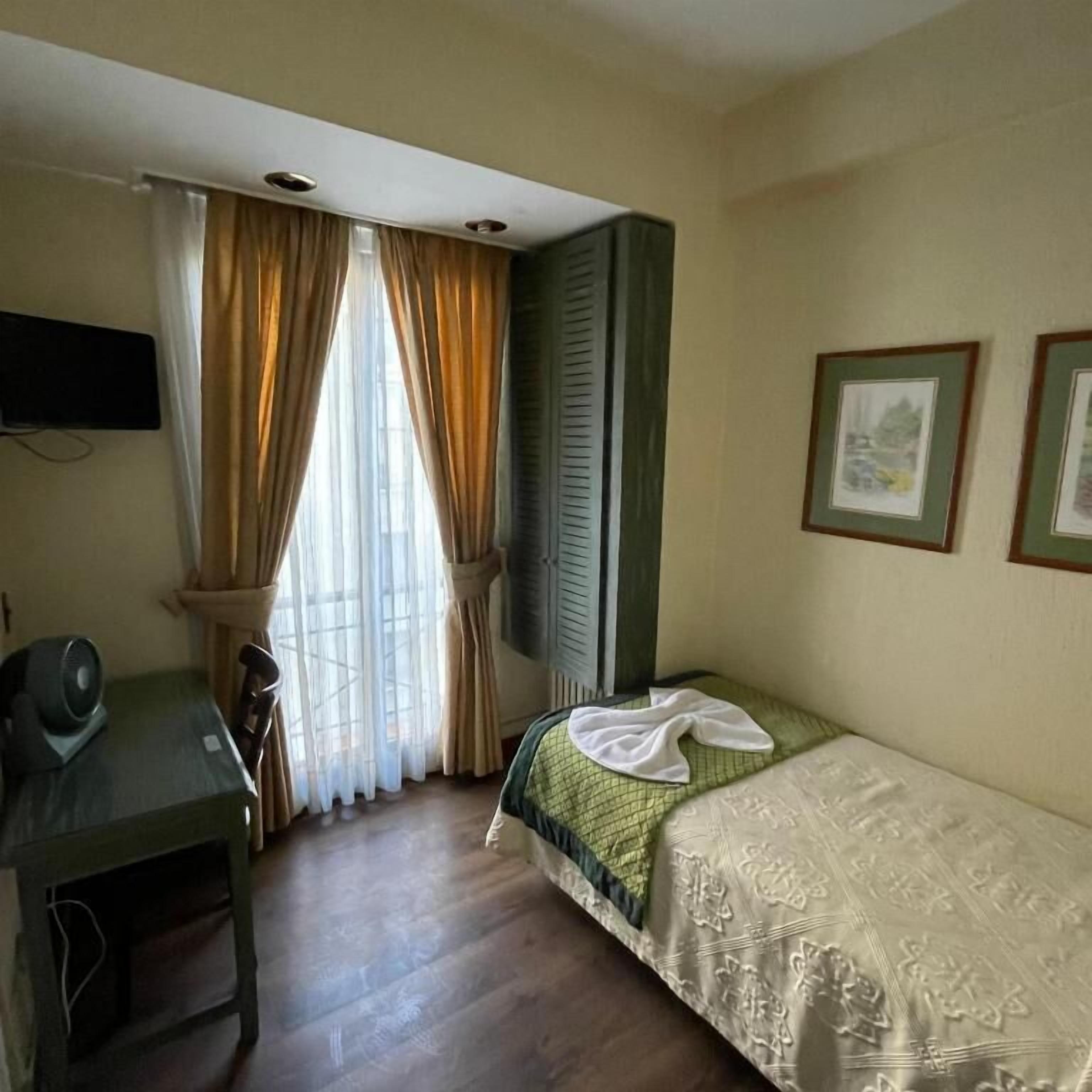 A small, simple room with a desk, a bed, and two framed pictures on the wall.