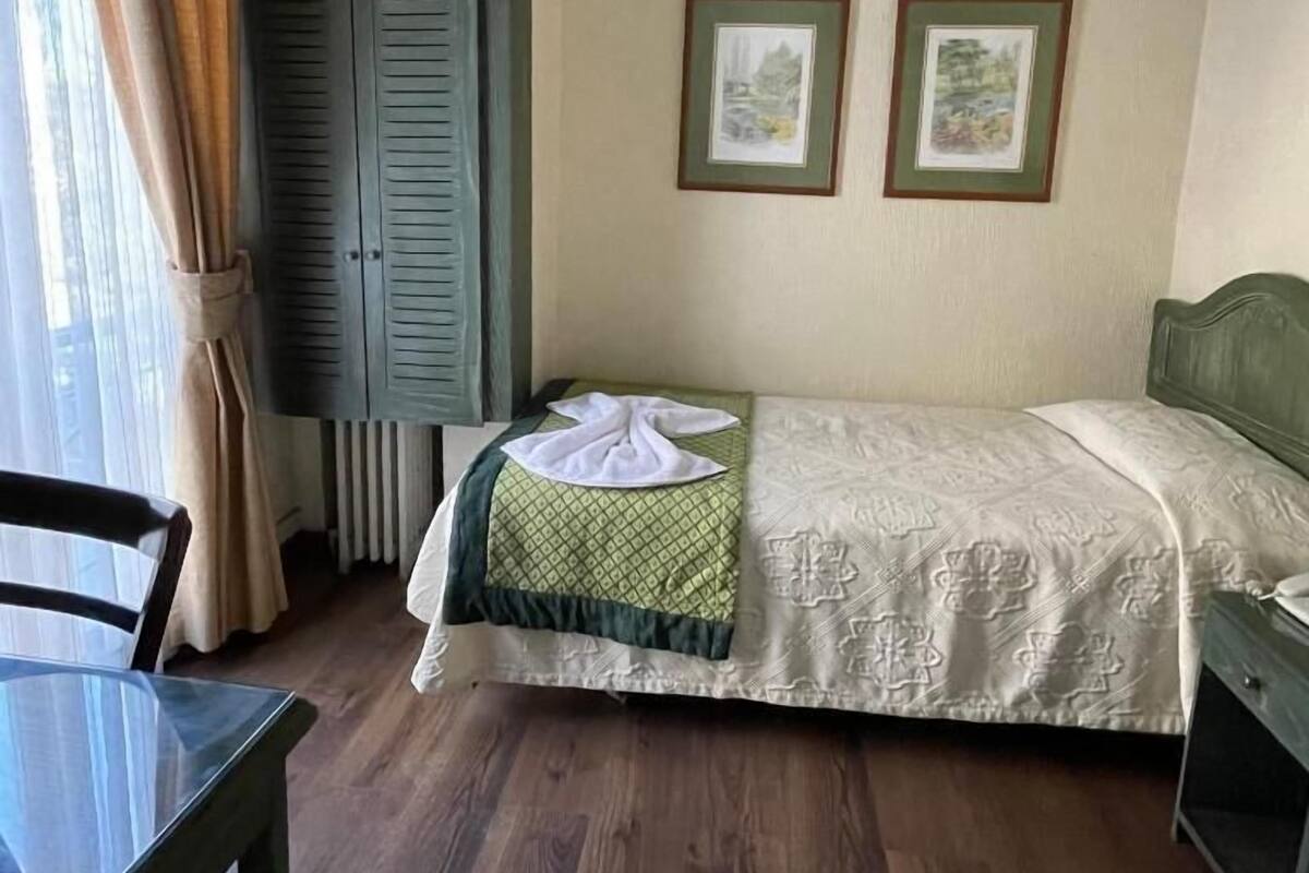 A small, neatly arranged bedroom with a single bed, a chair, and a table. There are two framed pictures on the wall, and a window with curtains.