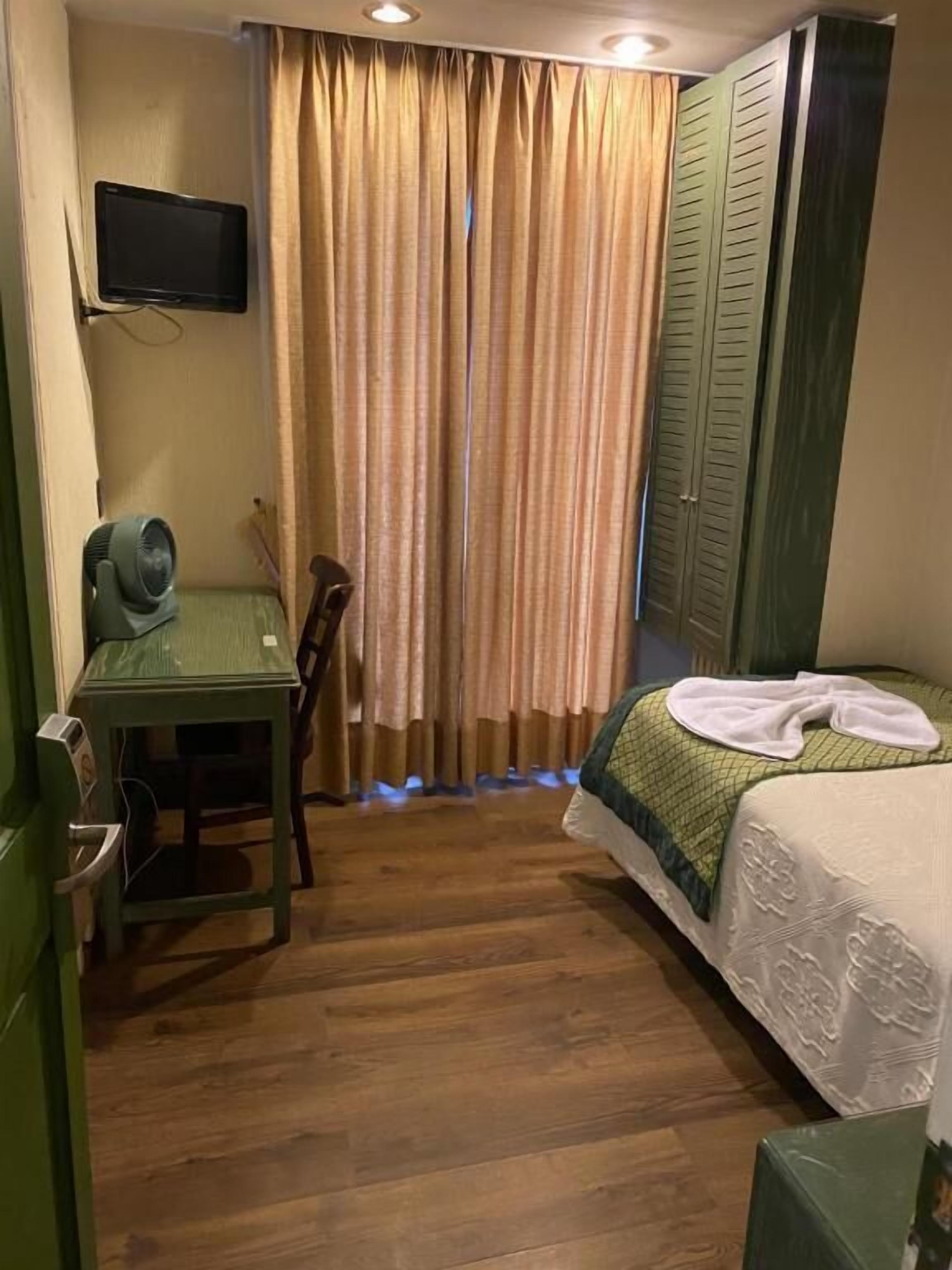 Habitació d'hotel amb un llit, una taula de treball, una cadira, un televisor i una finestra amb cortines.
