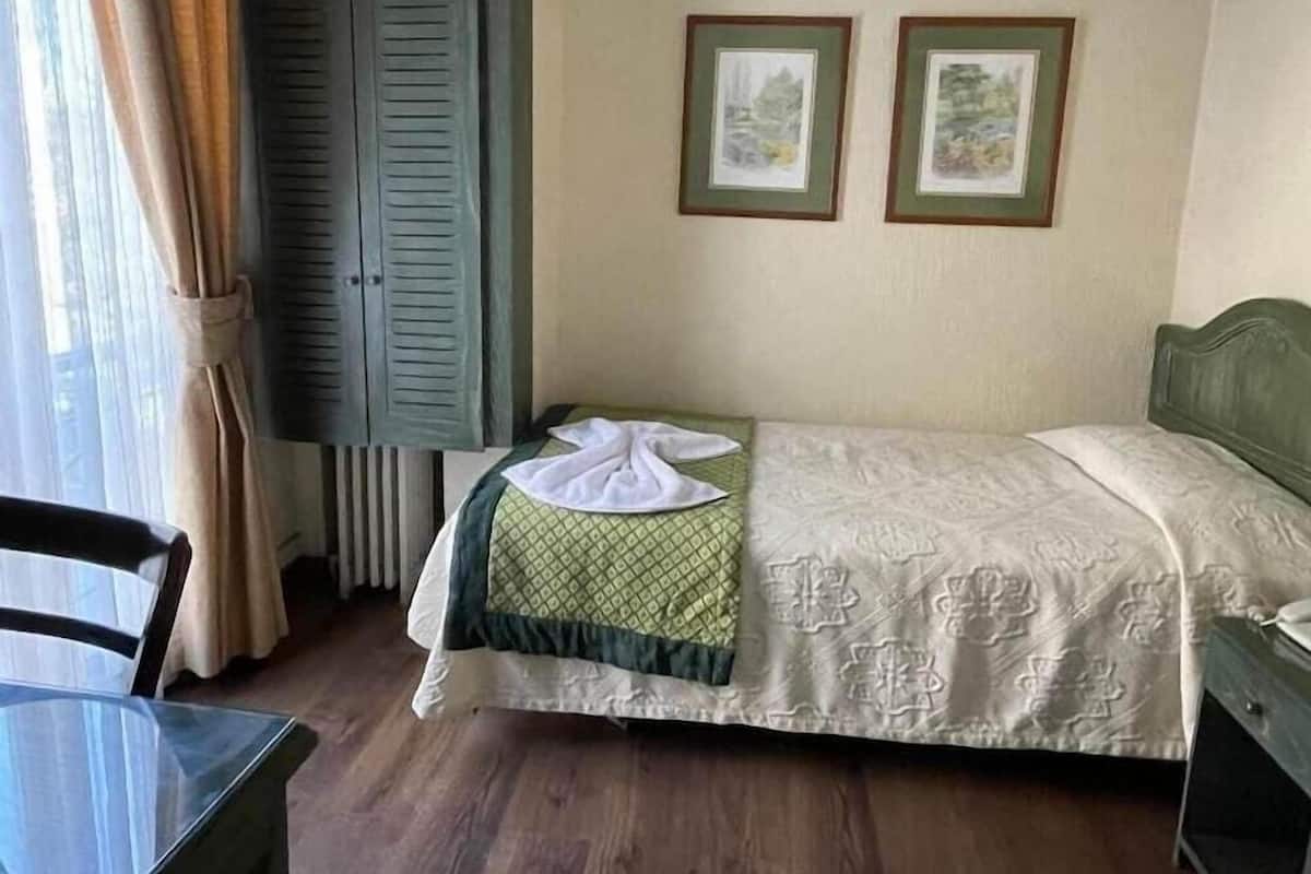 A small, neatly arranged bedroom with a single bed, a chair, a table, and two framed pictures on the wall.