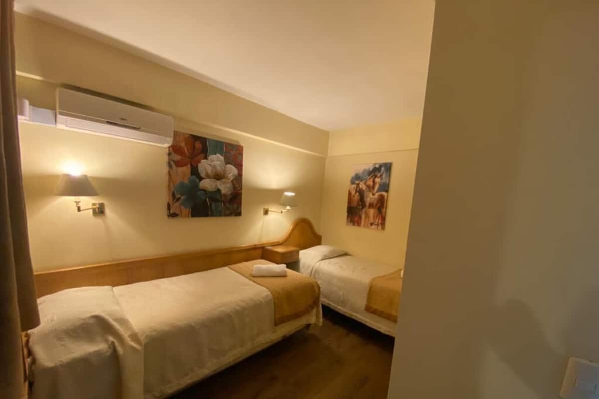A hotel room with two single beds, a wall-mounted air conditioner, and two wall-mounted lamps.
