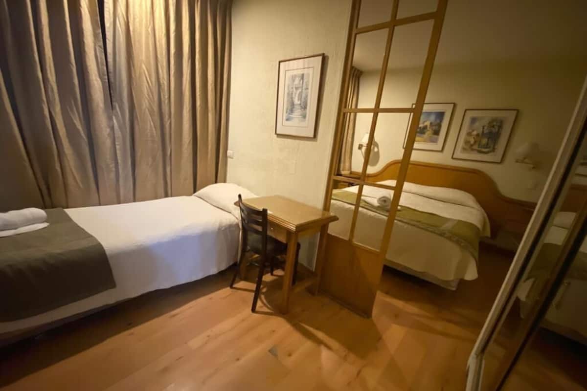 A hotel room with a bed, a desk, and a chair. There are curtains on the window and a painting on the wall.