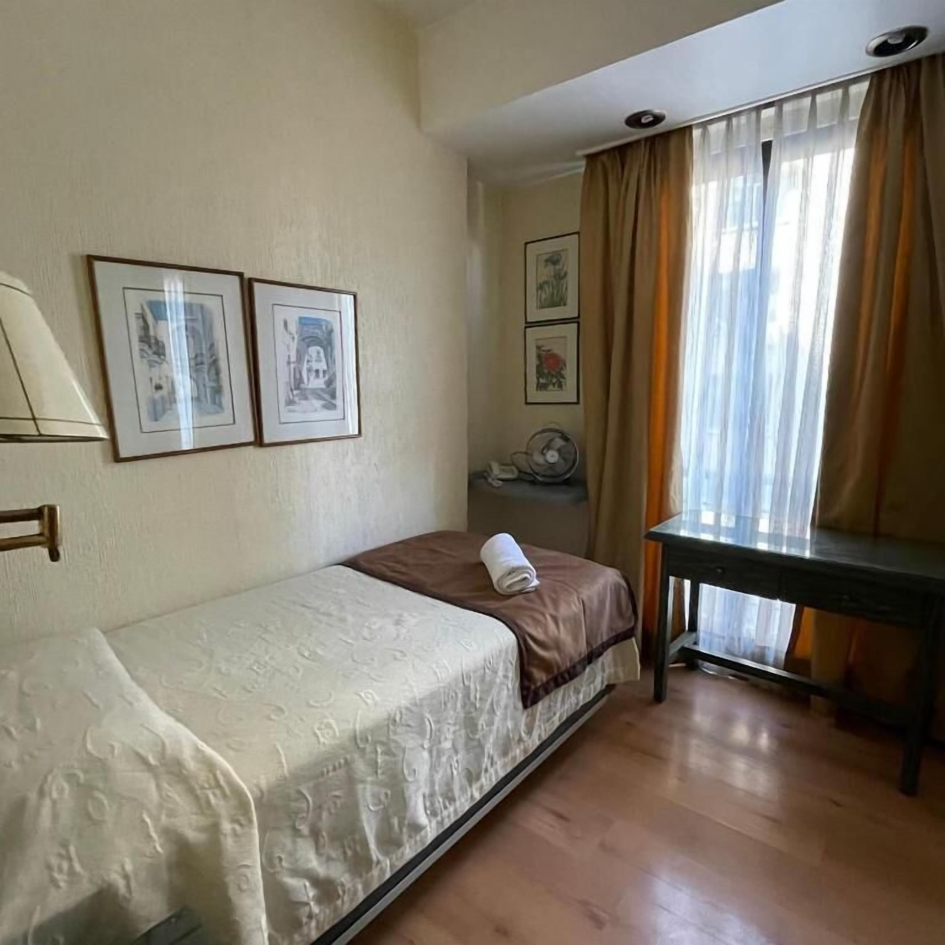 A hotel room with a bed, a desk, and a fan. There are three framed pictures on the wall.