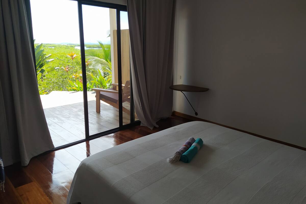 Une chambre d’hôtel avec un grand lit, une vue sur un jardin luxuriant grâce à une porte-fenêtre coulissante, et une petite table près de la porte.