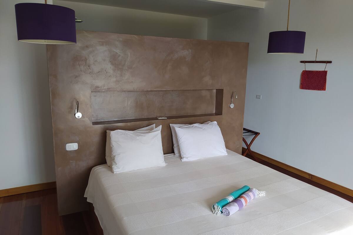 Une chambre d’hôtel avec un grand lit, une tête de lit, deux oreillers blancs, une couverture et une serviette enroulée posée sur le lit.