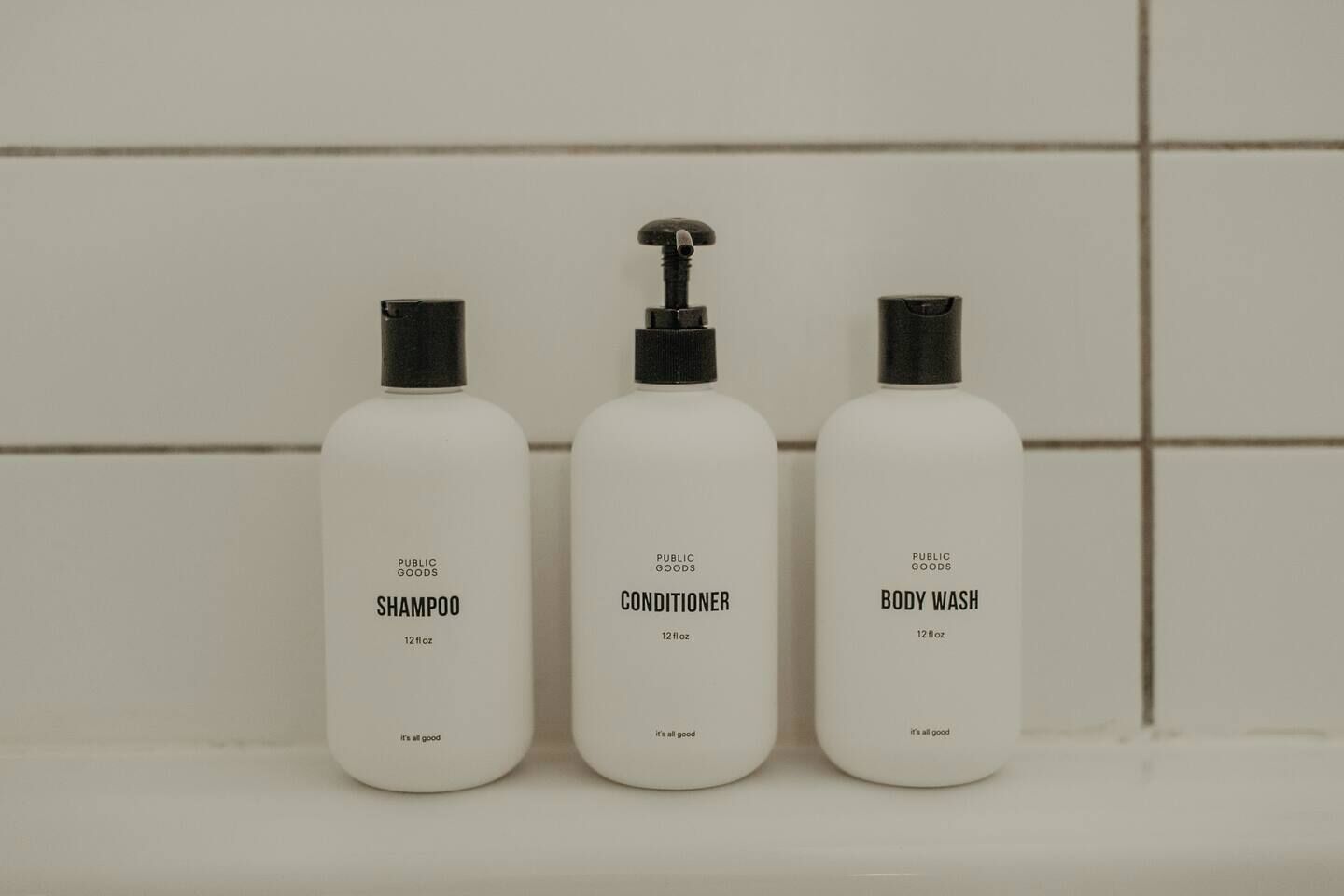 Tre flaconi di prodotti Public Goods: shampoo, balsamo e bagnoschiuma, ognuno con una capacità di 350 ml, su una superficie piastrellata.