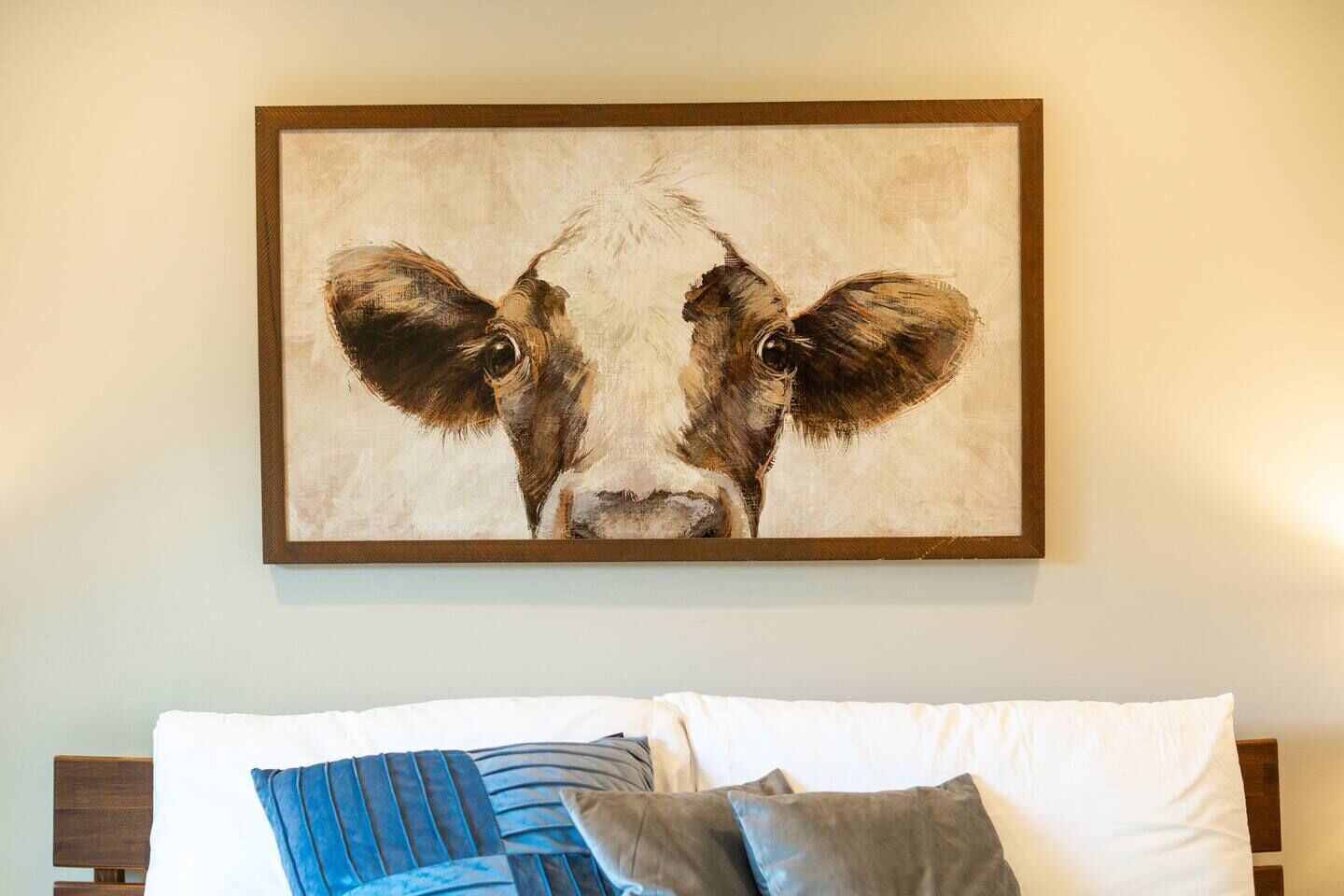 Un cuadro enmarco con el rostro de una vaca colgado sobre una cama con almohadillas blancas y azules.