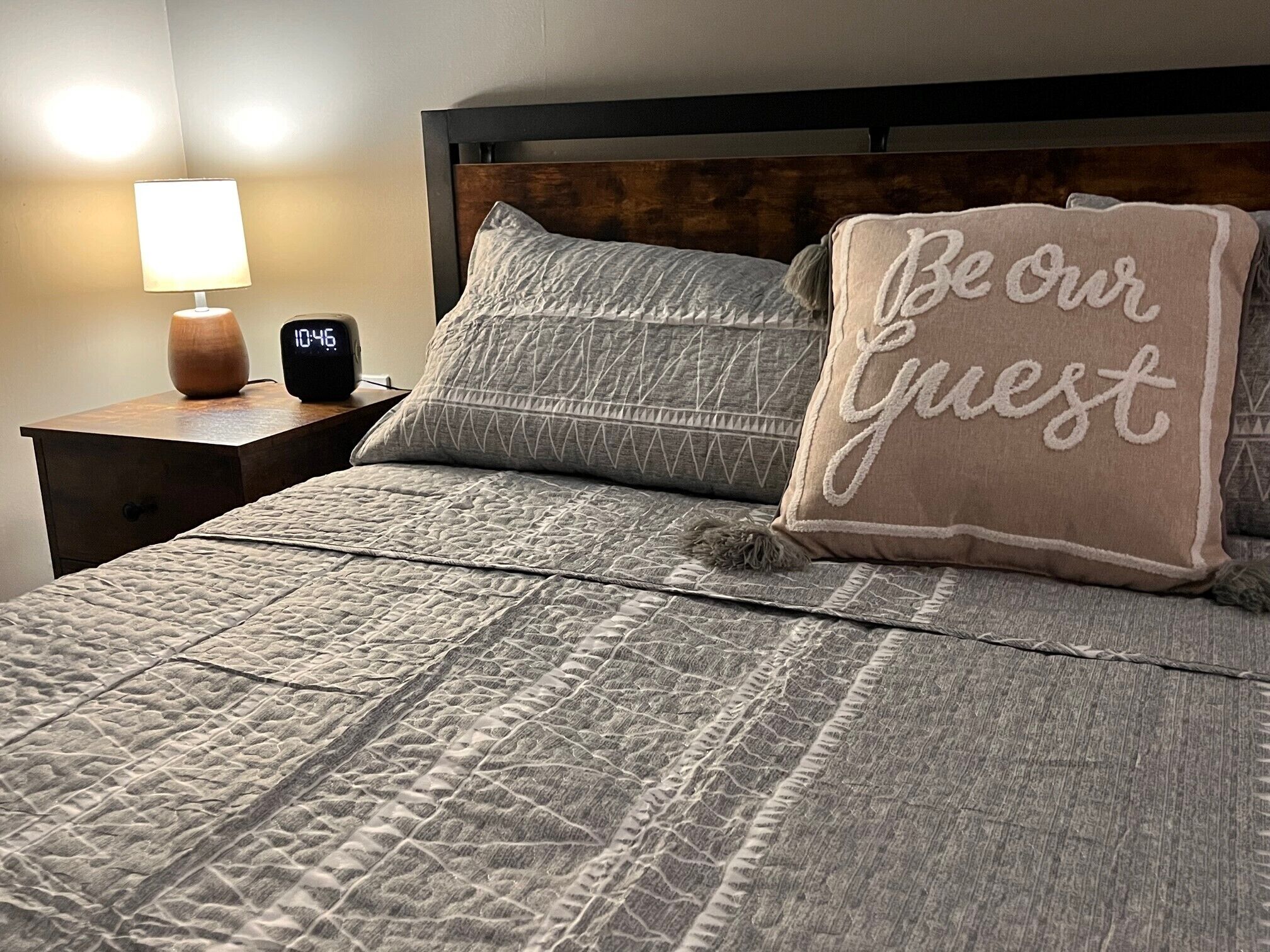 Un lit bien fait, avec une couvre-lit matelassée et des oreillers, une lampe de chevet et une horloge numérique affichant l’heure.