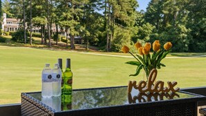 Une table en verre avec des bouteilles, un vase rempli de fleurs jaunes et une pancarte « détente » sur un balcon donnant sur un terrain de golf.