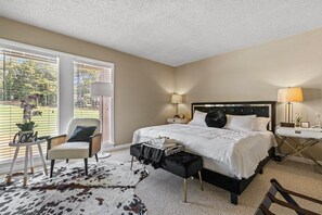 Un dormitorio con cama, mesitas de noche, lámpara, silla, una pequeña mesa con una planta y un tapete de cuero de vaca.