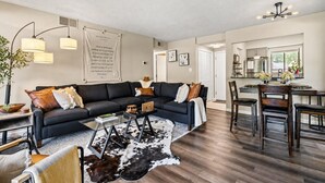 Un salon moderne avec un canapé d’angle, une table basse et un coin repas comprenant des chaises, et une cuisine en arrière-plan.