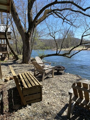 Eine Holzterrasse am Seeufer mit Adirondack-Stühlen und einer Feuerstelle, umgeben von Bäumen und freiem Blick auf den See.