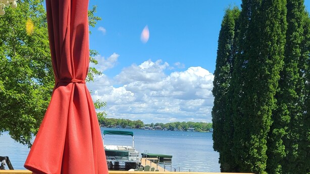 A table with a red umbrella, a view of a lake, and a wooden deck with chairs.