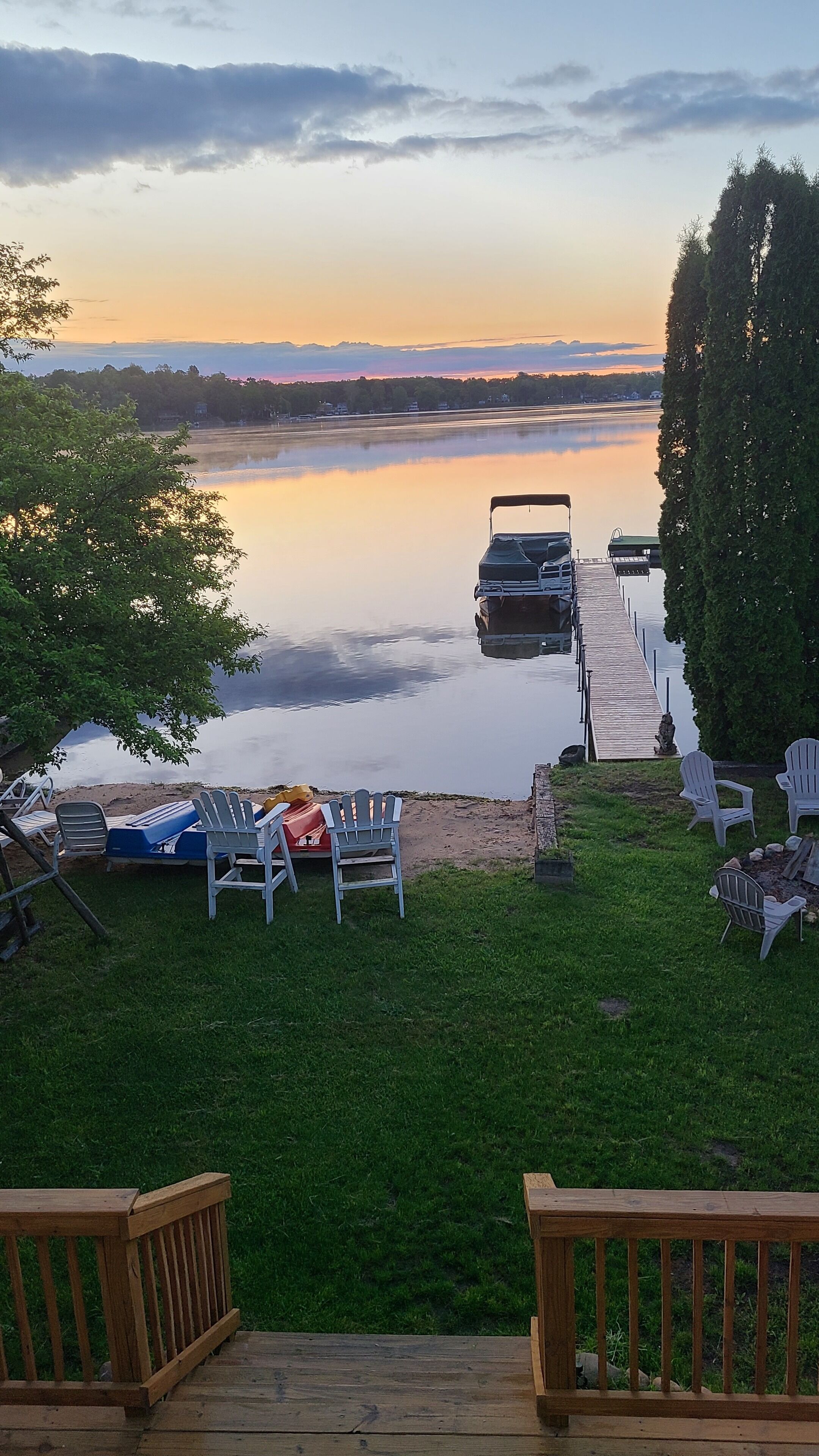 A lakeside deck with a dock extending into the water, surrounded by greenery and a sunset view.