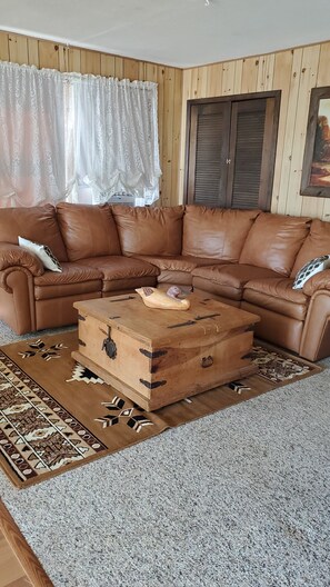 A corner living room with a brown leather sectional sofa, a wooden chest of drawers, patterned rugs, and wooden panel walls.