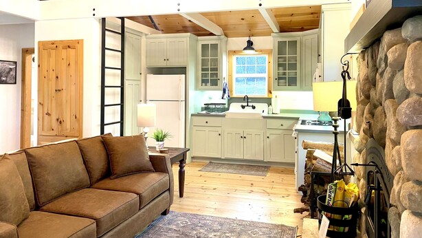A spacious living area with a brown sofa, wooden flooring, a kitchen with white cabinets, and a stone fireplace. - Duck Pond Cabin-Private Cabin on 400 Acre Ranch (Smartsville)
