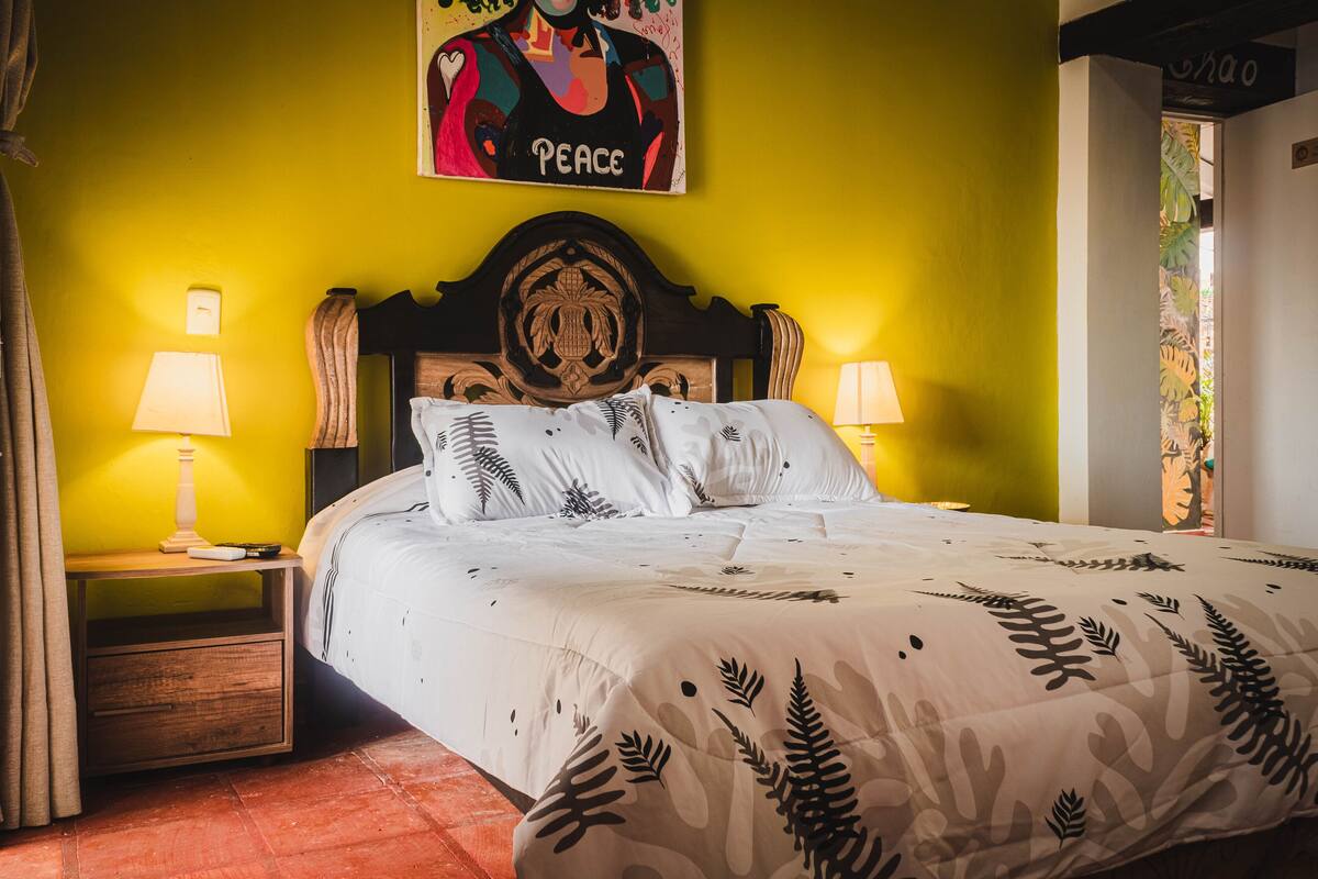 A bed with a patterned bedspread, a wooden headboard, two bedside lamps, and a wall with a 'Peace' poster.