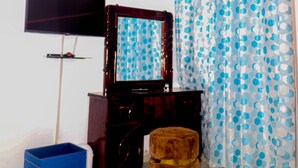 Un dormitorio con televisor, cómoda, espejo, caja azul para guardar objetos, taburete redondo y alfombra estampada.