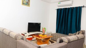 Una sala de estar con un sofá, un televisor, una pequeña mesa de madera con un jarrón y una cortina azul.