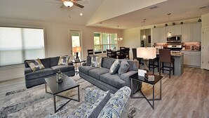 Un salon spacieux avec un canapé gris, une table basse et un pouf à motifs. On aperçoit à l’arrière-plan un coin repas avec une table et des chaises.