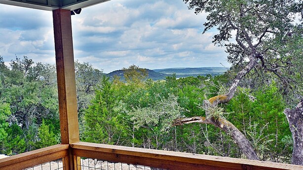 Un balcon en bois, doté d’une balustrade en métal, offre une vue sur une forêt dense et des collines lointaines.