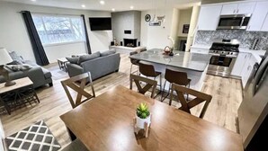 Una cocina moderna con una isla central, un comedor con mesa y sillas de madera, un sofá gris y un televisor montado en la pared.