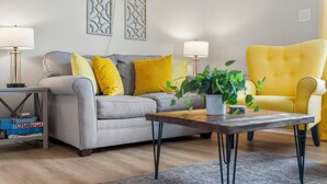 Een woonkamer met een grijze bank, gele kussens, een gele fauteuil, twee vloerlampen en een houten salontafel met een plant erop.