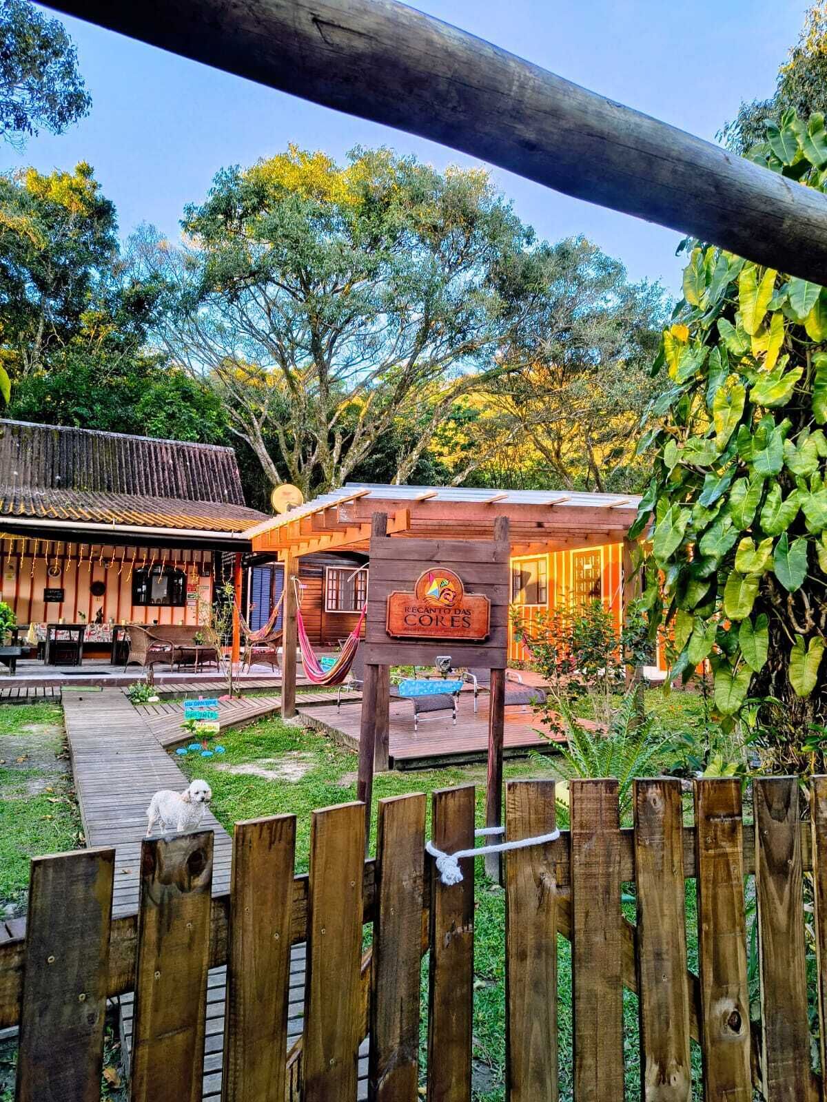 Un panneau en bois avec un logo et du texte, un hamac, une terrasse en bois et un espace clôturé où se trouve un chien.