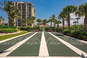 Un patio con canchas de shuffleboard, palmeras y un edificio de hotel al fondo.