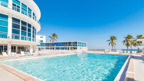 A modern hotel with a large pool, palm trees, and a clear blue sky.