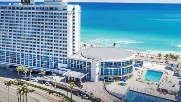 A large hotel with a central circular building, a pool, and a beach view.