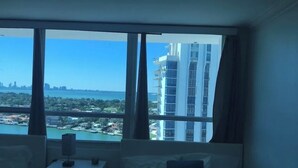 A hotel room with a bed, a bedside table, and a large window offering a view of a cityscape and water.