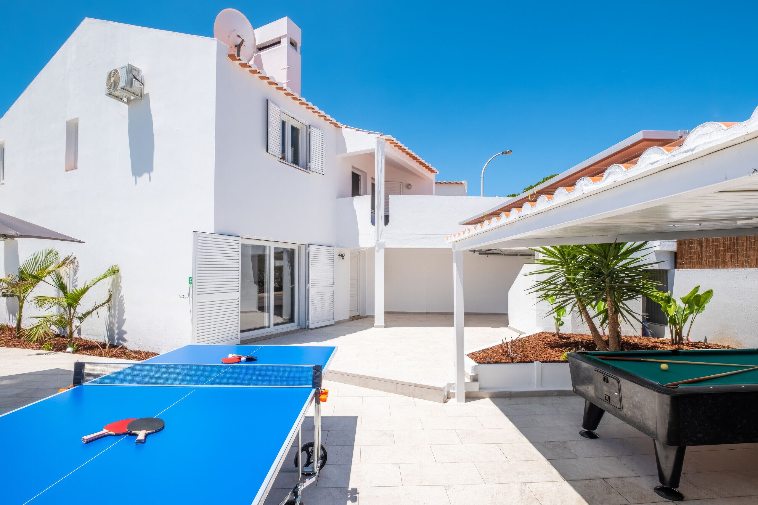 Une cour avec une table de ping-pong, un billard et un bâtiment aux murs blancs et au toit recouvert de tuiles.